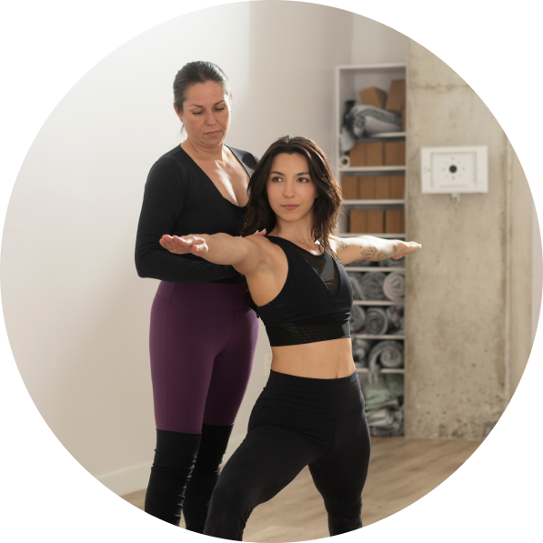 Two women yogis are standing in a warrior 2 yoga pose. One women is assisting the other with proper form.